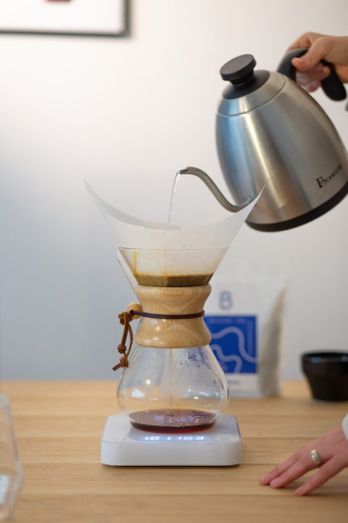 An electric kettle pouring hot water over specialty coffee grounds in a Chemex pour-over coffee brewing device.